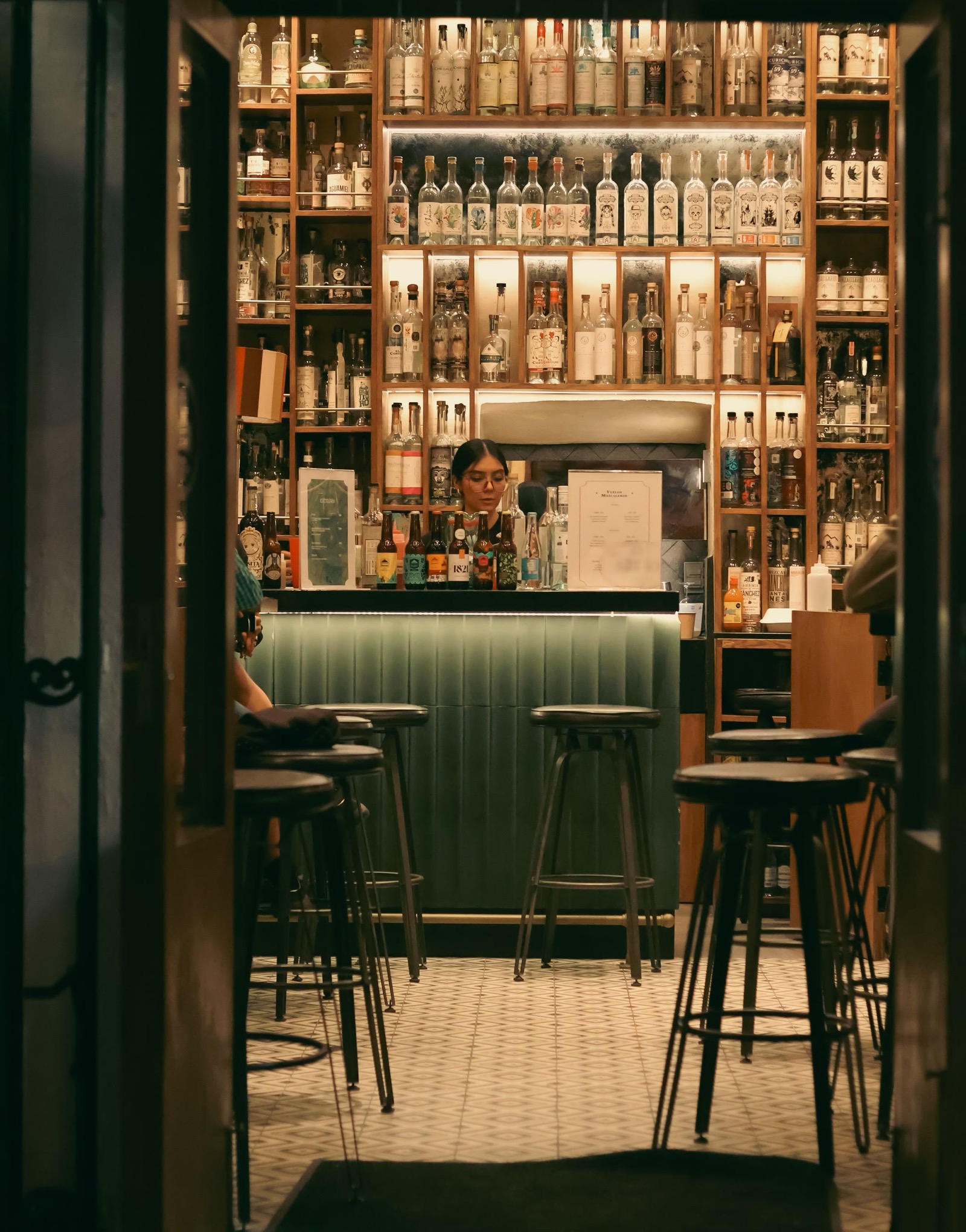 De noche, a través de una puerta abierta: una pared de repisas de madera llenas de botellas de mezcal iluminadas, una barra verde y bancos altos sobre piso de mosaico.
