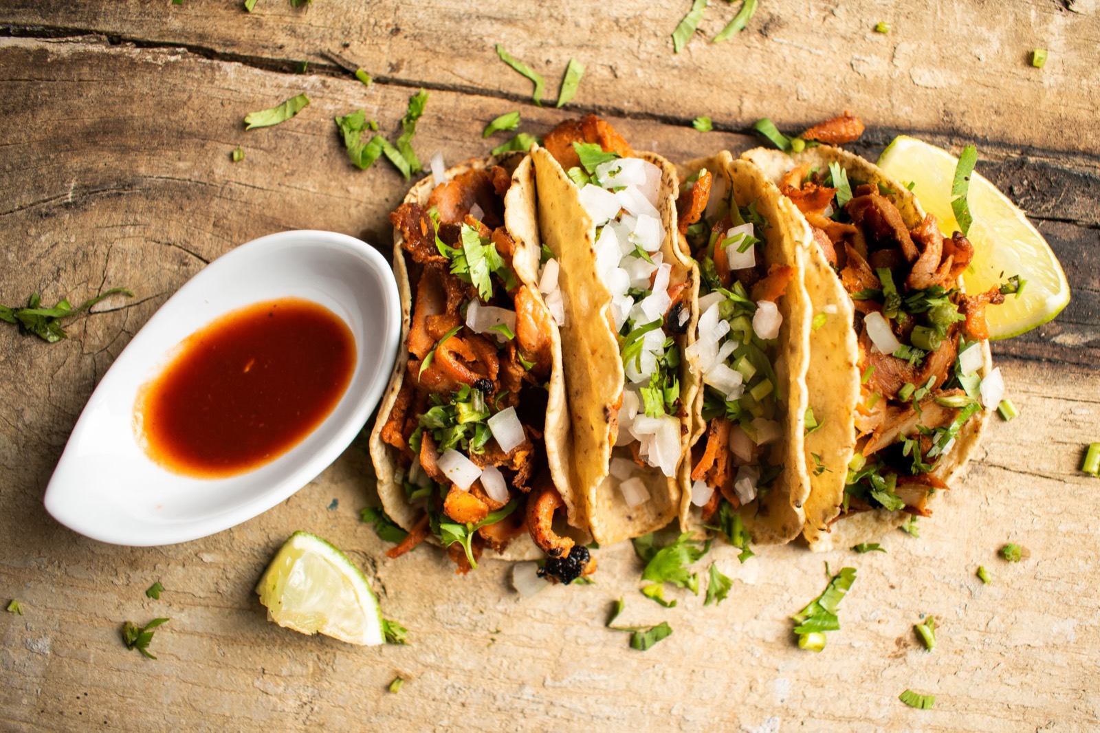 Cuatro tacos al pastor con cebolla picada y cilantro sobre una tabla de madera, con salsa roja y gajos de limón.