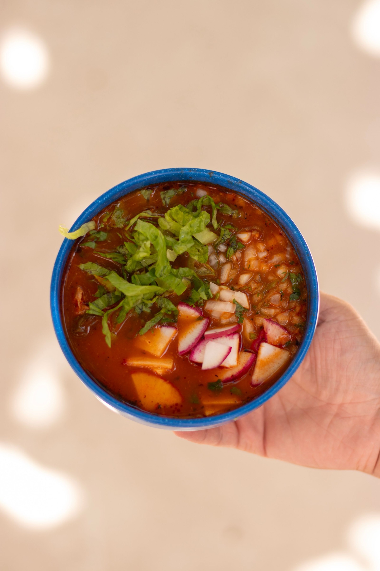 Un tazón azul de pozole rojo con maíz pozolero, lechuga, rábano y cebolla, sostenido en una mano.