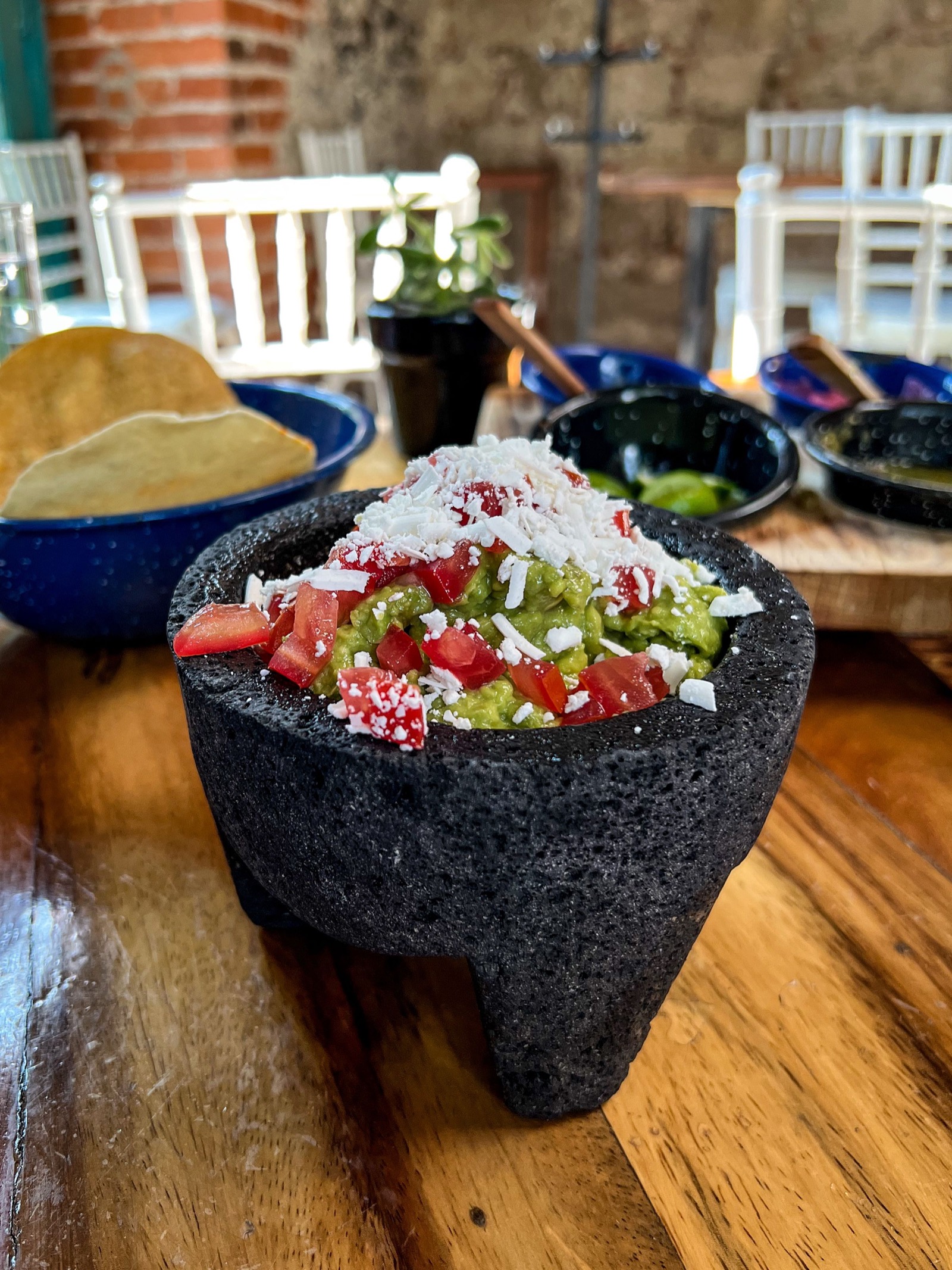Guacamole con jitomate y queso en un molcajete de piedra; detrás, tostadas y salsas en platos de peltre azul.
