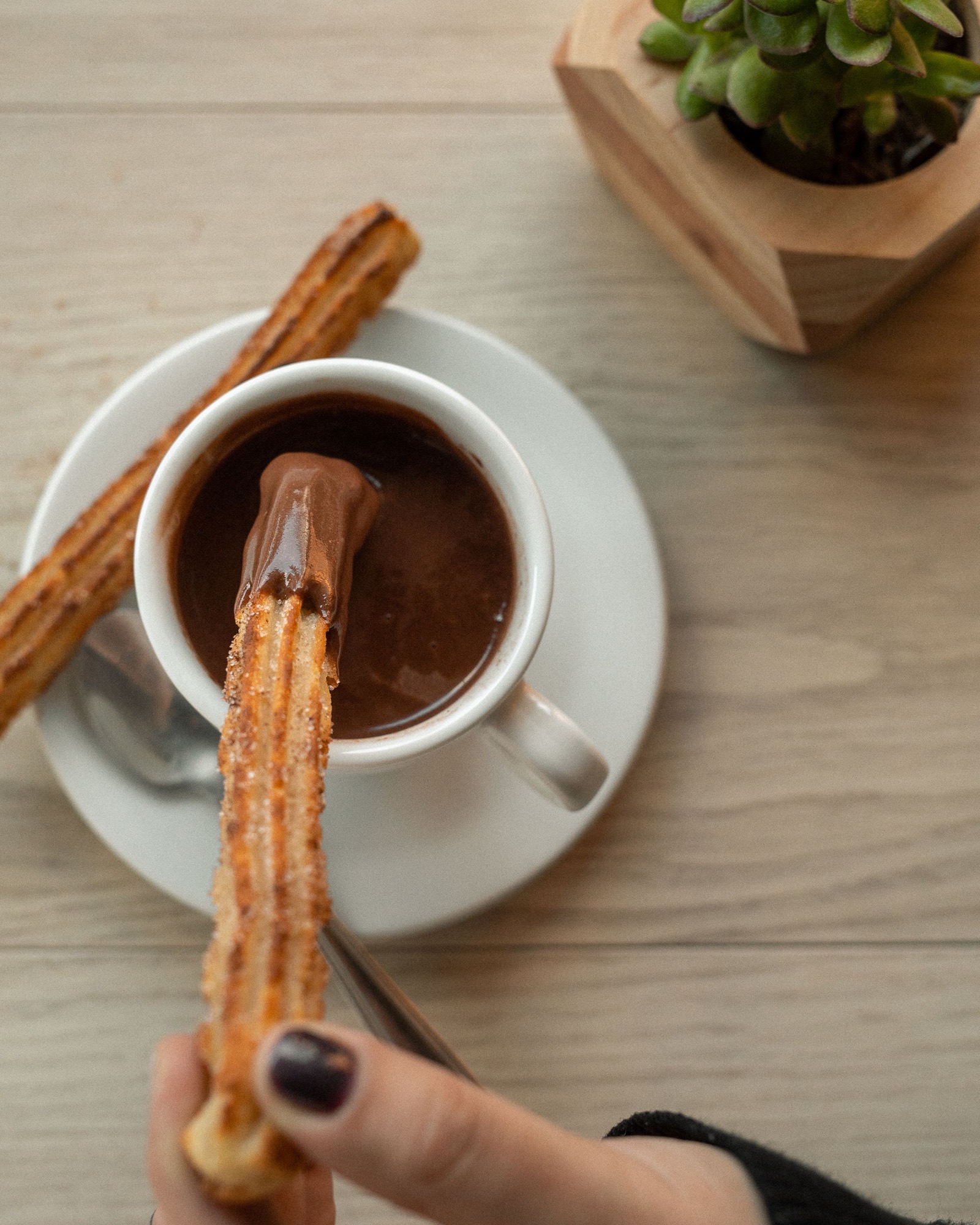 Una mano moja un churro en una taza de chocolate caliente; otro churro descansa en el plato.