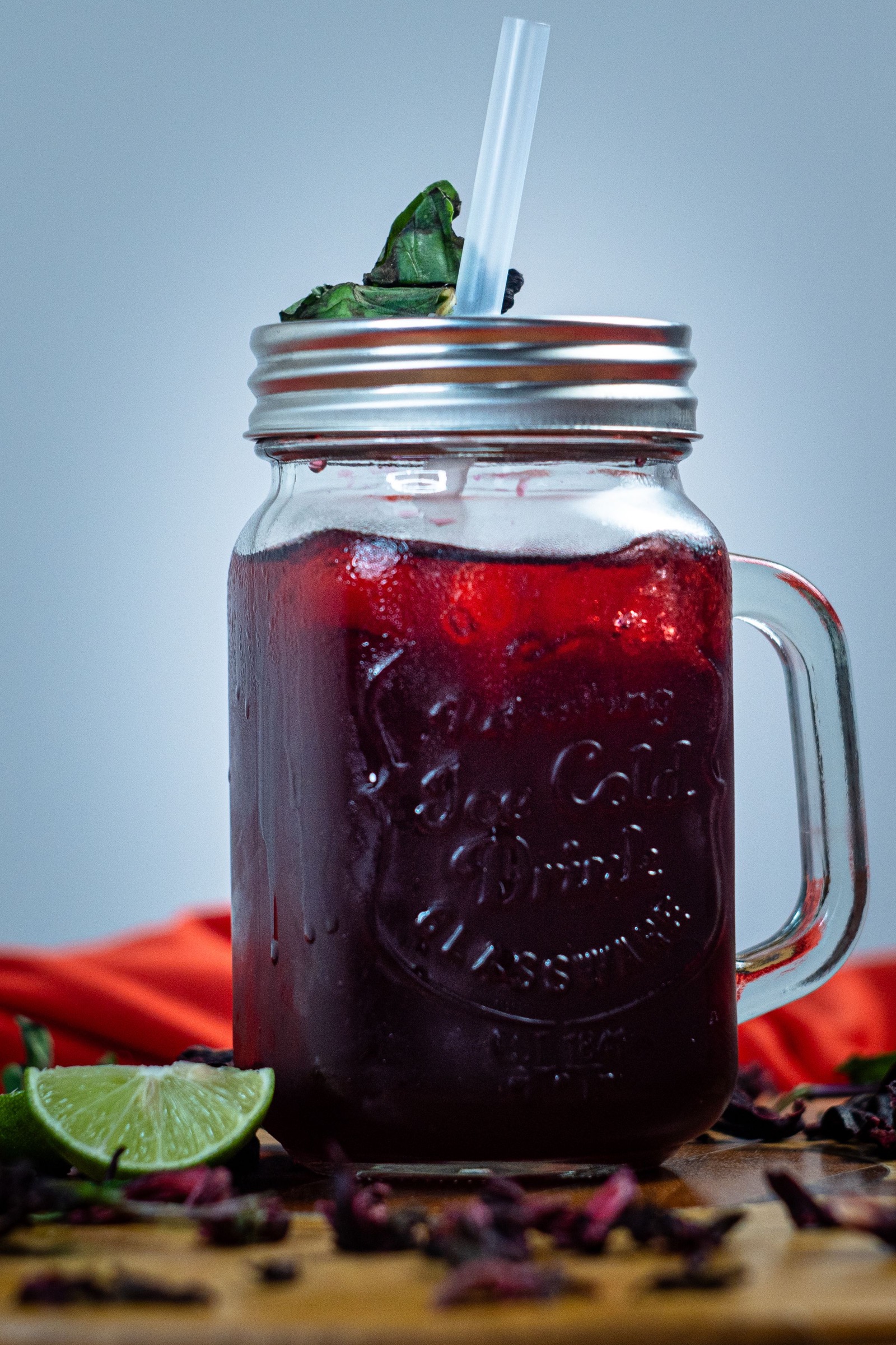 Agua de jamaica en un tarro de vidrio con popote, junto a flores de jamaica secas y un limón.