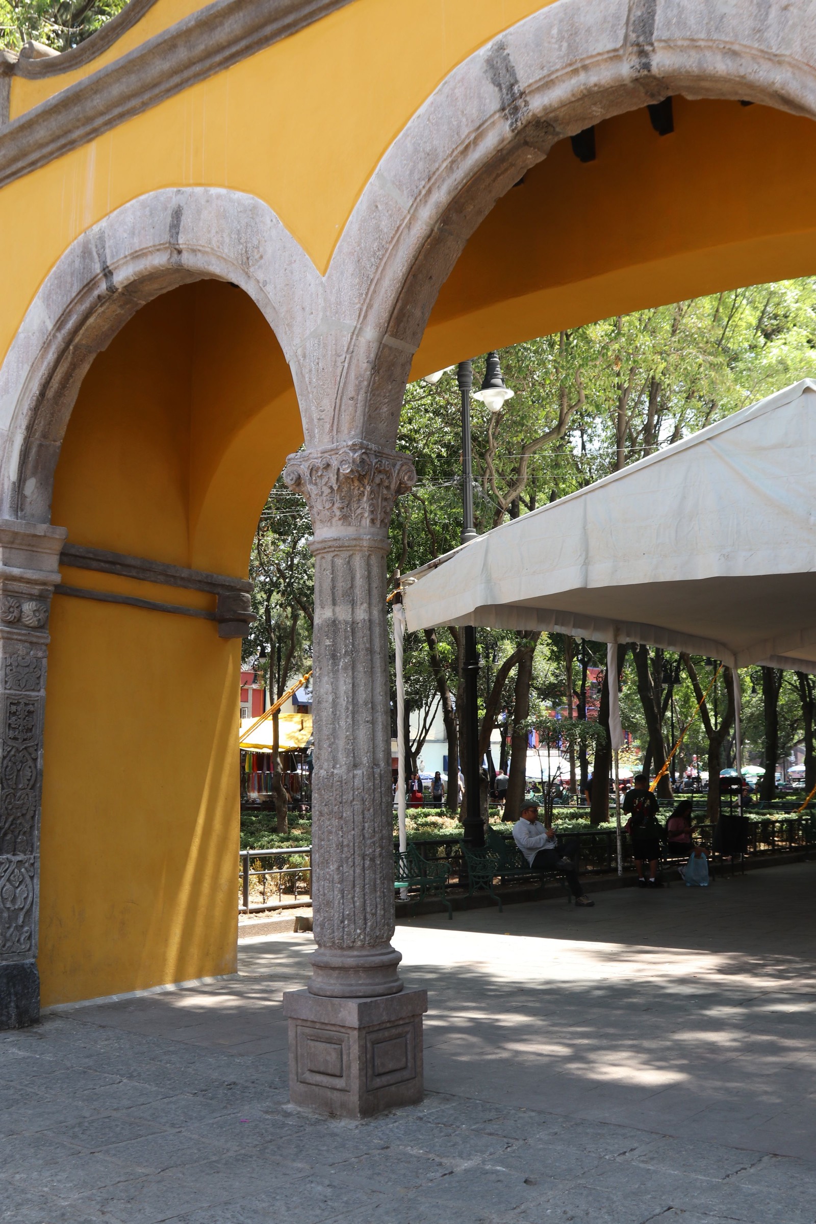 Arcos de cantera con columnas de piedra sobre un muro amarillo; al fondo, una plaza con árboles, bancas y un toldo blanco.